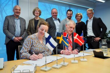 A group of people standing and sitting by a desk and watching a document being signed.